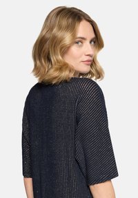 Femme blonde aux cheveux ondulés mi-longs portant un haut texturé bleu marine, regardant par-dessus son épaule sur un fond blanc.