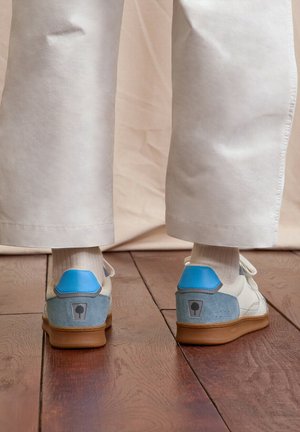 Person wearing white pants and white sneakers with blue accents, standing on polished wooden floor, viewed from behind.