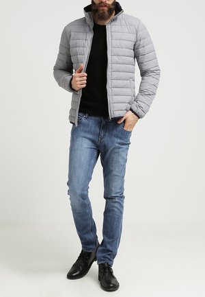Man med ljusgrå vadderad jacka, svart tröja, blå jeans och svarta läderskor, står med en hand i fickan.