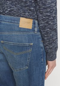 Personne portant un jean bleu Esprit et un pull bleu foncé à motifs, montrant la poche arrière et le patch en cuir de la marque.