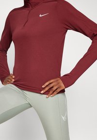 Bordowy długi rękaw sportowy top z wysokim kołnierzem i srebrnym logo Nike, zestawiony z jasnoniebieskimi legginsami z subtelnym graficznym akcentem.