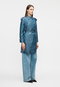 Γυναίκα όρθια που φοράει μπλε παλτό καμπαρντίνας με μοτίβο και ζώνη, ανοιχτό μπλε τζιν με φαρδιά μπατζάκια και μαύρα μυτερά παπούτσια, σε λευκό φόντο.