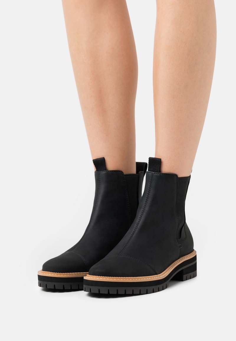 Bottines noires avec semelles épaisses à crampons et bordures beige, portées sur des jambes nues, debout sur fond blanc.