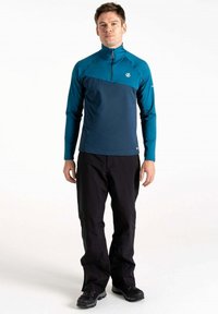 Türkis und dunkelblauses Langarm-Zipshirt mit hohem Kragen und Logo, kombiniert mit schwarzen Hosen und schwarzen Schuhen.