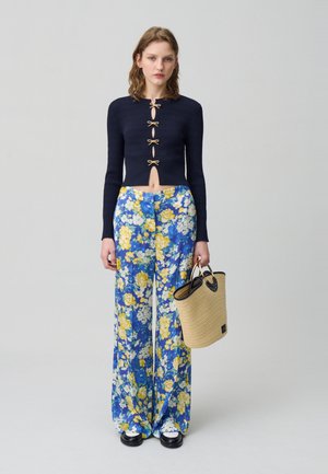 Top corto a coste blu navy con dettagli a fiocco dorati; pantaloni larghi floreali in blu, giallo e bianco; tiene in mano una borsa di paglia intrecciata.