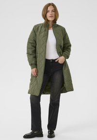 Olivgrön puffad långkappa med hög krage, dragkedjestängning och sidofickor. Matchad med svarta jeans och platta skor.
