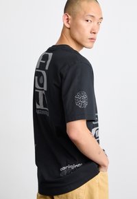 Svart bomulls-T-shirt med stor grå grafik på ryggen och en mindre logotyp på ärmen. Korta ärmar, avslappnad passform och rund hals.