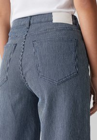 Les jeans en denim rayés présentent une taille haute, des poches arrière et une étiquette en cuir blanc au niveau de la taille. Les rayures sont bleu marine et blanches.