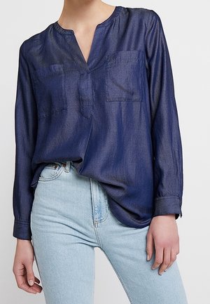 Frau trägt eine dunkelblaue Bluse mit langen Ärmeln und zwei Brusttaschen, in hellblaue hoch taillierte Jeans gesteckt.
