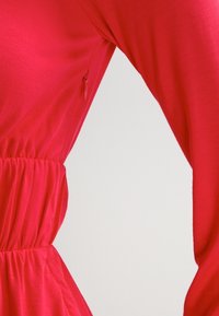 Robe rouge avec une taille cintrée, des manches longues et un tissu doux et lisse. Présente des fronces élastiques à la taille et une fermeture éclair latérale.