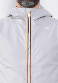 Giacca leggera grigia chiara con chiusura frontale a zip. Presenta una striscia multicolore al centro e un cappuccio. Texture liscia e lucida.