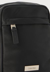 Sort nylon taske med en glat tekstur, der har lynlås-lukning, sølvfarvet hardware og en guld logo-plakette. Justerbar stropdetalje.