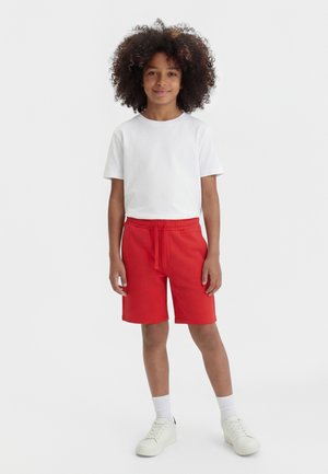 Barn med krøllete hår iført hvit t-skjorte, røde shorts, hvite sokker og hvite joggesko som står mot en ensfarget bakgrunn.