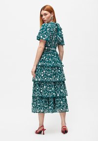 Teal kukka-teemainen kerrostettu mekko lyhyillä hihoilla, valkoisilla kuvioilla jalaisella yksityiskohdalla selässä; yhdistetty punaisiin korkokenkiin.
