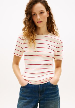 Ung kvinde med krøllet hår iført hvid T-shirt med røde vandrette striber og blå jeans, hænderne i lommerne, neutralt baggrund.