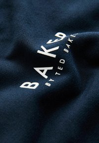 Marineblauem Stoff mit leicht strukturiertem Finish, mit weißer Aufschrift "BAKED BY TED BAKER" in moderner Schriftart, teilweise zerknittert.