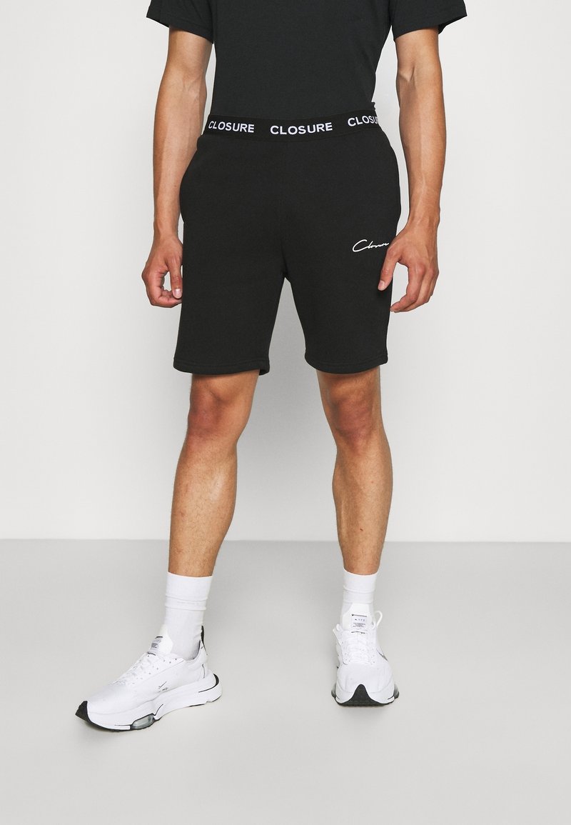 Shorts noirs pour hommes avec taille élastique imprimée "CLOSURE", mélange doux de coton, coupe ajustée, et petit logo blanc sur la jambe inférieure gauche. Portés avec des baskets blanches.