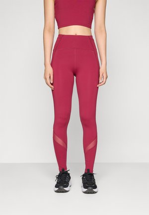 Leggings bordeaux taille haute avec une texture lisse, des panneaux en maille au niveau des mollets et une coupe ajustée, associés à des chaussures de sport noires.