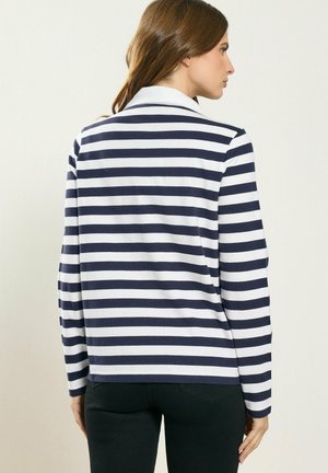 Vrouw met bruin haar die een wit en donkerblauw horizontaal gestreept shirt met lange mouwen en een zwarte broek draagt, zijwaarts kijkend tegen een effen achtergrond.