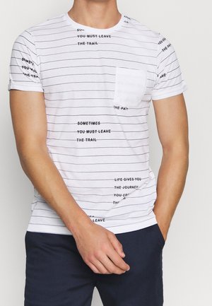 Camiseta blanca de manga corta con finas rayas horizontales negras y frases de texto negras dispersas, usada por una persona con una mano en el bolsillo.