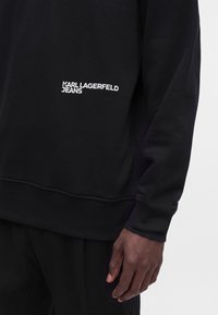 Μαύρο φούτερ με λευκό λογότυπο "Karl Lagerfeld Jeans" στο στήθος, φορεμένο από άτομο με σκουρόχρωμο δέρμα, ορατό από το μέσο του κορμού έως το χέρι.