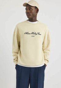 Sudadera color crema con texto bordado en negro "Nautica Sailing Crew 1983", combinada con pantalones azul oscuro y una gorra clara a juego.