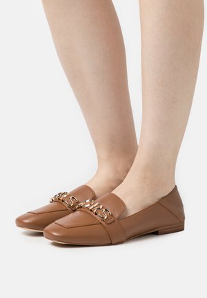 Chaussures plates en cuir marron avec une chaîne décorative dorée portant l'inscription "MARC JACOBS", portées sur des pieds sur fond blanc.