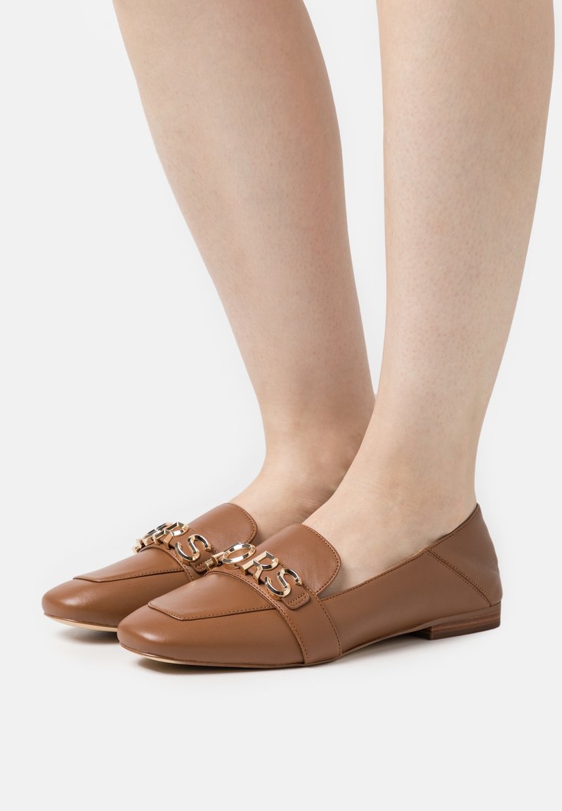 Chaussures plates en cuir marron avec une chaîne décorative dorée portant l'inscription "MARC JACOBS", portées sur des pieds sur fond blanc.