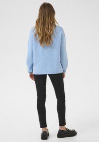 Světle modrý oversize svetr vyrobený z měkké tkaniny, spárovaný s černými slim-fit džíny a černými platformovými botami, zachycený ze zadní strany.