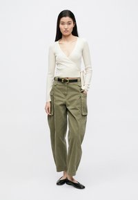 Pulover înfășurat bej deschis cu textură cu dungi; pantaloni cargo verde măsliniu cu buzunare laterale; curea neagră și pantofi negri cu talpă plată.