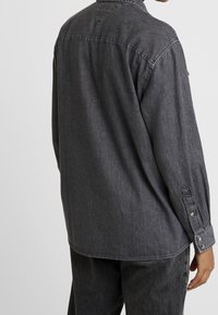Camicia in denim grigio antracite con maniche lunghe, polsini con bottoni e vestibilità rilassata. Presenta un colletto classico e una leggera texture.