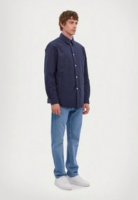 Mann steht in einer dunkelblauen knöpfen Jacket, hellblauen Jeans und weißen Sneakern vor einem schlichten hellen Hintergrund.