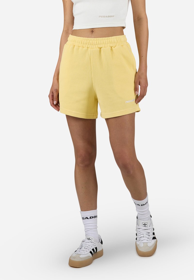 Shorts gialli a coste con una vita elastica, abbinati a calze bianche e scarpe da ginnastica bicolori in bianco e nero. Decorati con un dettaglio del logo.