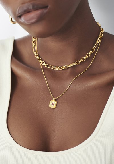 Colares em camadas de ouro, apresentando uma corrente grossa e uma corrente delicada com um pendente retangular e uma pedra preciosa incrustada, usados com uma blusa branca.