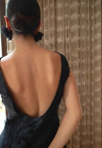 Musta mekko, jossa on syvä V-malli selkäpuolella, tekstuuritettu kangas ja ohuet olkaimet. Hiukset suihdeltu nutturalle mustilla kukkaaksonteilla.