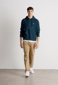 Felpa teal con cordini, tasca a marsupio, pantaloni beige e sneakers bianche. Il modello è in piedi su una superficie liscia di colore grigio chiaro, contro un muro bianco.