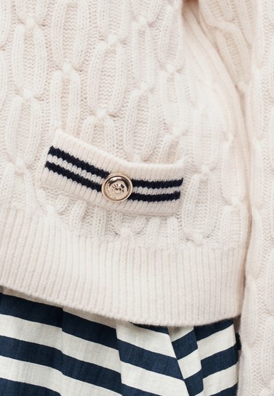 Hvid kabelstrikket sweater med ribbet kant og dekorativt stribet flap med rund sølvknap over et navyblåt og hvidstribet stof.
