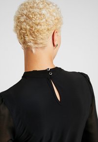 Person mit kurzen, lockigen blonden Haaren, die ein schwarzes Oberteil mit einem Träne-ausschnitt und zwei kleinen Knopfverschlüssen am Kragen hinten trägt.