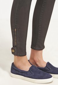 Jeans skinny neri con zip dettagliata alle caviglie, abbinati a scarpe slip-on blu navy con una finitura in suede testurizzato.