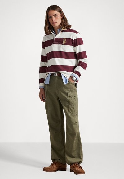 Tânăr cu părul până la umeri, purtând un tricou de rugby cu dungi, o cămașă albastră dedesubt, pantaloni cargo de culoare măslinie și cizme maro, stând în picioare.