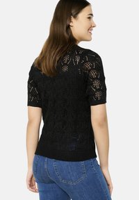 Juodas megztas viršus su trumpomis rankovėmis, puošnus lapų raštu, tekstūrinė medžiaga ir surištas apvadas, derinamas su mėlynais džinsais.