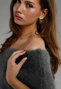 Grå strikket sweater med en fuzzy tekstur, båret uden om skulderen. En models hånd holder blidt på stoffet og fremviser hendes guldøreringe med bølge mønster.