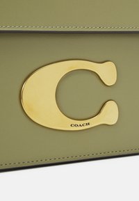 Maslinasta kožna torbica s velikim zlatnim C-oblika logotipom i utisnutim detaljem "COACH". Šavovi duž rubova su vidljivi.
