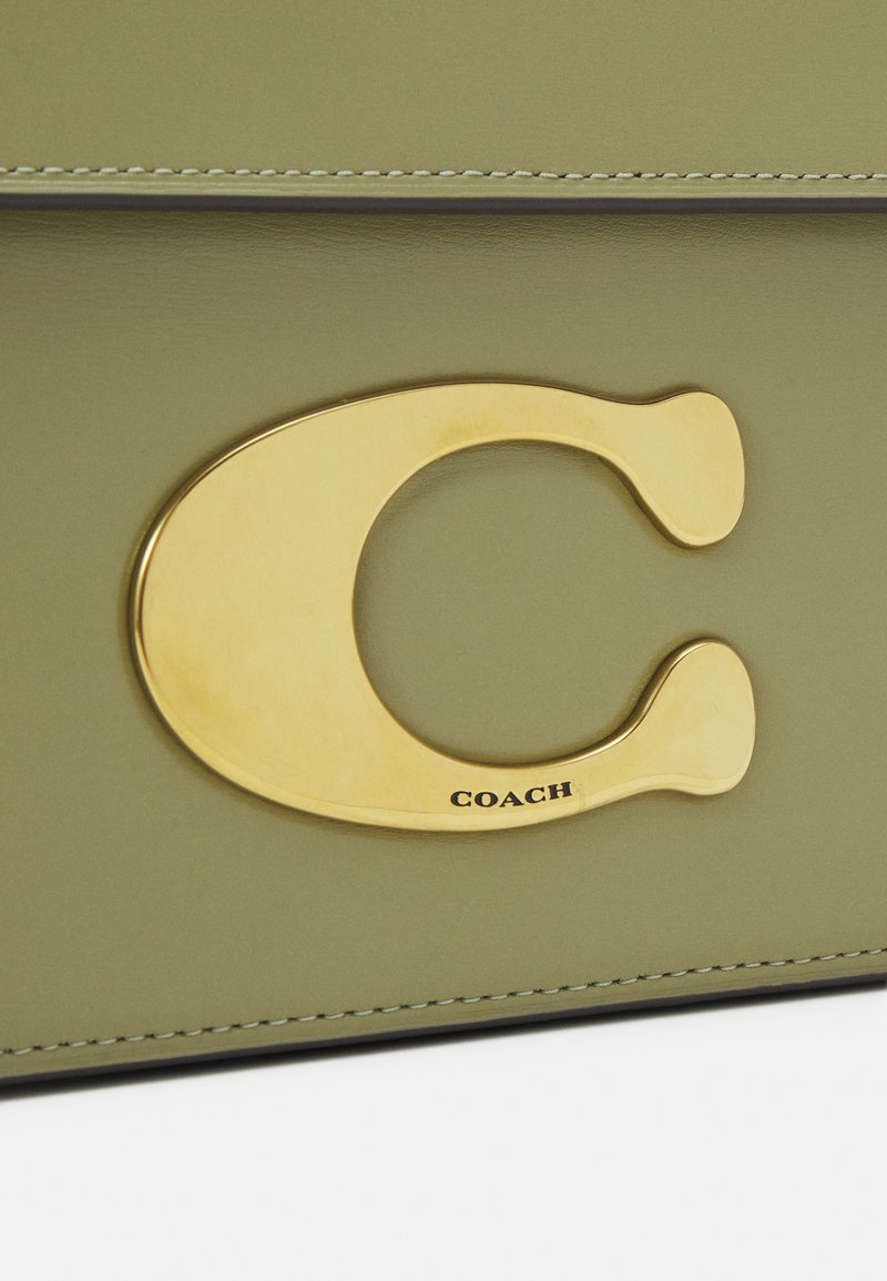 Olivengrüner Lederhandtasche mit einem großen goldfarbenen C-förmigen Logo und geprägtem "COACH"-Detail. Die Nähte entlang der Kanten sind sichtbar.
