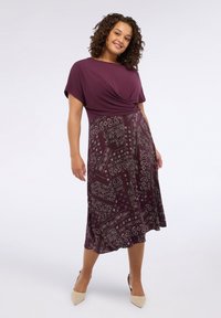 Femeie îmbrăcată într-o rochie purpurie cu un top solid și o fustă cu model paisley, stând și zâmbind pe un fundal alb simplu.