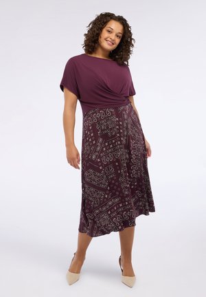 Frau in einem lila Kleid mit einem einfarbigen Oberteil und einem Rock im Paisleymuster, die steht und lächelt vor einem schlichten weißen Hintergrund.