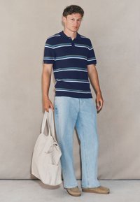 Tricou polo cu dungi bleumarin, cu mâneci scurte, combinat cu blugi albastru deschis și pantofi maro, ținând o geantă tote bej cu bretele.