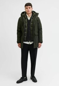 Grüne gepolsterte Winterjacke mit Kapuze, kombiniert mit einem schwarzen Strickpullover, einem weißen Hemd und schwarzen Hosen. Schwarze Schuhe runden das Outfit ab.