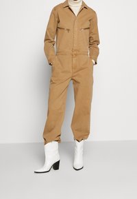 Mono de algodón color marrón claro con mangas largas, bolsillos con cremallera en el pecho y bolsillos laterales, combinado con botas vaqueras blancas y una pieza para el cuello ajustada.