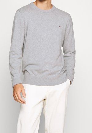 Muž nosící světle šedý svetr s dlouhým rukávem a malým logem na hrudi a bílé kalhoty, stojící s jednou rukou v kapse.